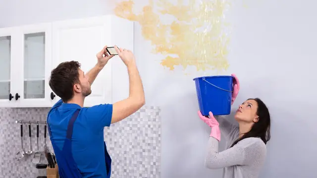 Man photographing a water-stained wall while a woman holds a bucket beneath the leak