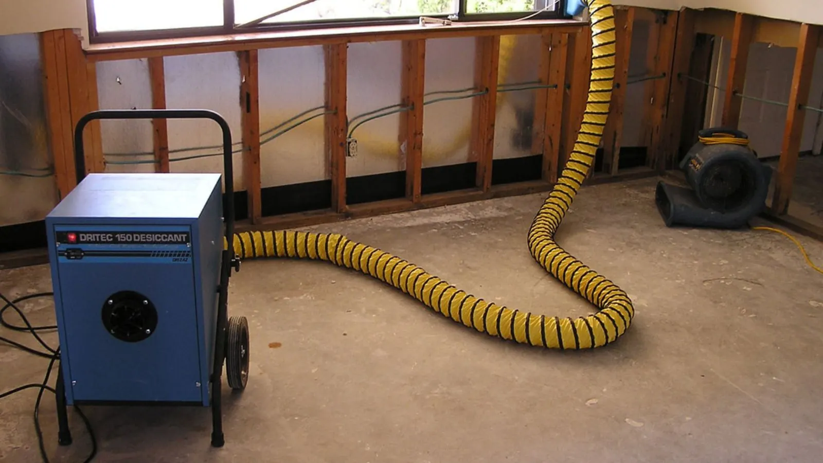 Dehumidifier with a yellow duct hose and air mover drying a room where wall sections were cut out after water damage